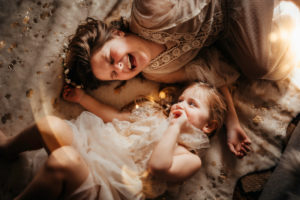 Family Photographer, a baby and her older sister admire each other, older sisters laughs, they lay in bed linens