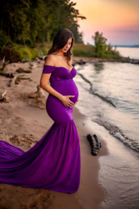 Maternity Photographer, a pregnant woman stands at the beach's shore in a dress at sunset